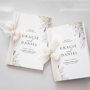 Può includere: Due programmi di nozze bianchi legati con nastri color crema. I programmi presentano i nomi "Gracie e Daniel" in elegante corsivo, insieme alla data e al luogo del matrimonio. Accenti floreali in viola e verde adornano gli angoli.
