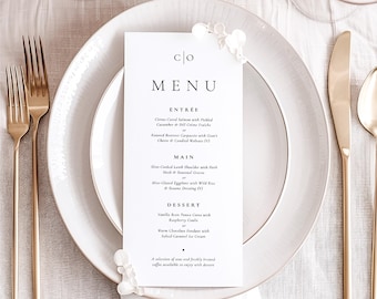 May include: A white menu card with the word "MENU" in black text. The menu lists the following items: Entrée, Main, and Dessert. The menu items are written in black text on a white background. The menu card is on a white table setting with gold silverware and a white plate.