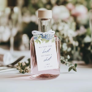 Könnte beinhalten: Kleine, transparente Glasflasche gefüllt mit rosa Flüssigkeit, mit hellbraunem Verschluss. Ein Etikett mit dem Text "WE TIED THE KNOT SO TAKE A SHOT" ist mit weißer Schnur und blauen floralen Akzenten am Hals befestigt. I&F 12.05.25 ist aufgedruckt.