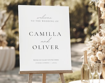May include: A white wedding welcome sign on a wooden easel. The sign reads "welcome to the wedding of Camilla and Oliver." A champagne bottle and two glasses are on a table in the foreground. The sign also includes the text "We're so glad you're here."