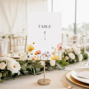 May include: A rectangular white table number card featuring the text TABLE 1 in black serif font, decorated with a colorful watercolor illustration of various wildflowers and a small bee at the bottom, held by a gold metal stand.