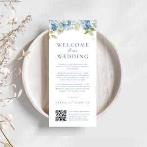Può includere: Un cartello di benvenuto per matrimoni bianco con accenti floreali blu e il testo "WELCOME to our WEDDING". Il cartello include un codice QR e i nomi Freya e Isabelle.