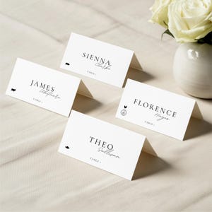 May include: Four white place cards with black text and small illustrations. Each card displays a name in elegant script, such as "Sienna Clarke" and "James Whitmore." A vase of white roses sits in the background.