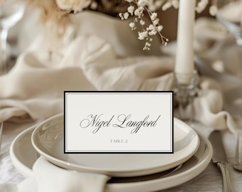 Pode incluir: Uma mesa formal com um cartão de nome com o nome "Nigel Langford" e "TABLE 2" em escrita elegante, colocado sobre um prato branco. O cartão tem uma borda preta. A configuração inclui um guardanapo dobrado e está sobre uma toalha de mesa branca.