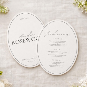 May include: Two cream-coloured oval wedding menu cards with black text. One says "Welcome Amelia Rosewood", the other "Food Menu". The menu lists entree, main, and dessert options. The date 21.12.2025 is also visible.