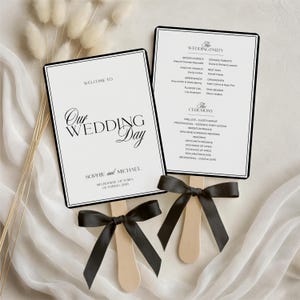 May include: Two wedding ceremony programs with black borders and black bows. The programs read "Welcome to Our Wedding Day" and list the wedding party and ceremony details. The programs are held by wooden sticks.