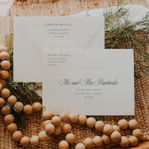 Puede incluir: Dos sobres color crema con texto negro. Uno dice "SOPHIE & MICHAEL" y detalles de la dirección. El otro dice "Mr and Mrs Pembroke" con una dirección. Los sobres están sobre una superficie tejida con cuentas de madera.