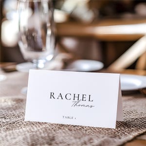 Pu&ograve; includere: Un segnaposto bianco con il nome "Rachel Thomas" stampato in nero, con "Table 1" sotto. Il biglietto &egrave; su una superficie strutturata di colore neutro, con uno sfondo sfocato di vetreria e stoviglie.