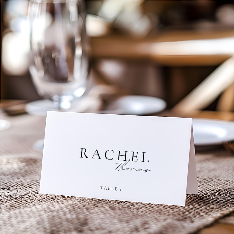 Pu&ograve; includere: Un segnaposto bianco con il nome "Rachel Thomas" stampato in nero, con "Table 1" sotto. Il biglietto &egrave; su una superficie strutturata di colore neutro, con uno sfondo sfocato di vetreria e stoviglie.