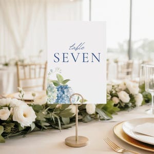 May include: A white table number card with the word "SEVEN" in navy blue, decorated with blue hydrangea flowers. The card is held by a gold metal stand, on a table with floral arrangements and gold-rimmed plates.