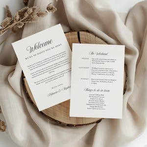 May include: Two cream-colored wedding invitation cards with elegant black script are displayed on a wooden slice. The left card reads "Welcome" and the right card lists the weekend's events. The cards are set against a neutral fabric background.