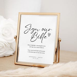 May include: A gold-framed sign with the text "Sign our Bible" in cursive script. The sign also includes instructions for guests to highlight a meaningful verse and sign their name beside it. The text "Your words and faith will become a cherished part of our love story" is also included, along with the couple's names and wedding date.