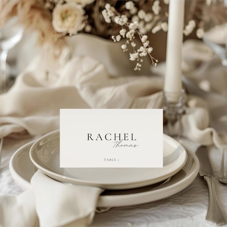 Pu&ograve; includere: Un'apparecchiatura formale con un piatto bianco, un tovagliolo piegato e un biglietto da visita. Il biglietto recita "RACHEL Thomas" e "Table 1". L'allestimento &egrave; su una tovaglia color crema, con una candela e una composizione floreale sullo sfondo.