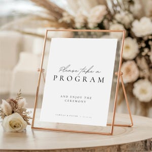 May include: A rose gold metal framed sign with the text "Please take a PROGRAM" and "AND ENJOY THE CEREMONY". The sign is on a wooden surface with floral arrangements in the background. The sign is for a wedding or special event.