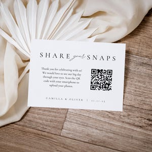 以下が含まれることがあります： 「SHARE your SNAPS」の文字とQRコードが記載された白いカード。カードには「ご参加いただきありがとうございます！」というテキストと、写真をアップロードするためのコードをスキャンする手順も含まれています。カードは木製の表面に置かれています。