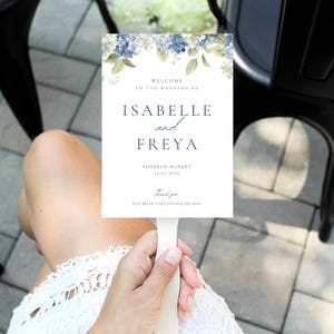 Può includere: Un cartello nuziale bianco con decorazioni floreali blu, tenuto da una persona. Il cartello recita "BENVENUTI AL MATRIMONIO DI ISABELLE e FREYA" e include la data 12.07.2025 e "Grazie per aver preso parte al nostro grande giorno."