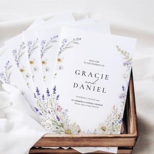 May include: A stack of white wedding programmes featuring a watercolour floral border of wildflowers in shades of purple, yellow, and green. The text reads: Welcome to the wedding of Gracie and Daniel, Saturday 21 August 2026, St Andrew's Catholic Church.