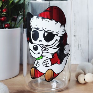 May include: A clear glass tumbler with a festive design featuring a cartoon character wearing a Santa hat and holding a snowman. The character has black eyes, a stitched mouth, and a red and white outfit.