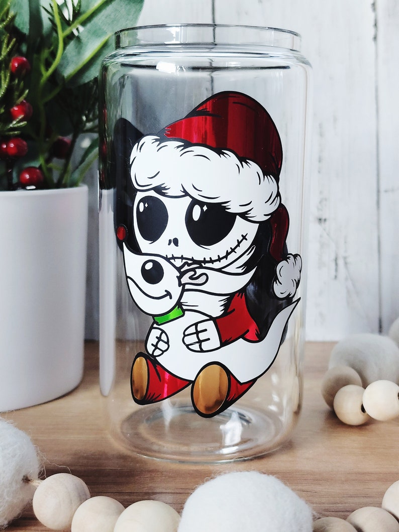 May include: A clear glass tumbler with a festive design featuring a cartoon character wearing a Santa hat and holding a snowman. The character has black eyes, a stitched mouth, and a red and white outfit.