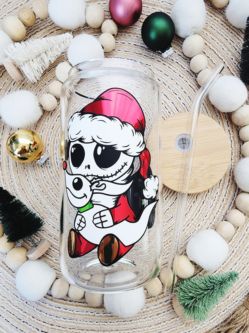 May include: A clear glass tumbler with a festive design featuring Jack Skellington from The Nightmare Before Christmas wearing a Santa hat and holding Zero. The tumbler has a red and white Santa hat design and a straw.