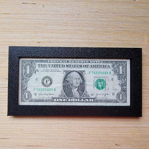 May include: A framed one-dollar bill. The bill features a portrait of George Washington and the text "ONE DOLLAR". The frame is black and rectangular, with a textured surface. The bill is centered within the frame.