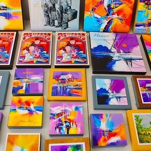 May include: A collection of framed abstract paintings in various colors, including blue, yellow, purple, and orange. Some of the paintings feature the word "Hawaii" and "Maui Bay".