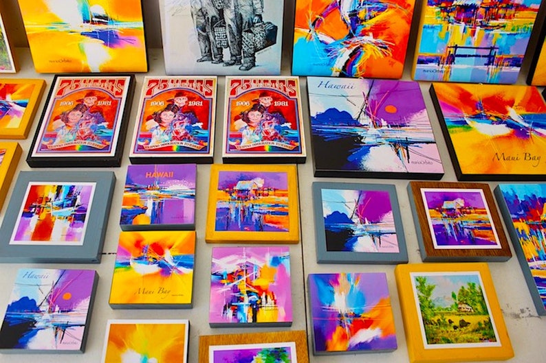 May include: A collection of framed abstract paintings in various colors, including blue, yellow, purple, and orange. Some of the paintings feature the word "Hawaii" and "Maui Bay".