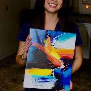 May include: A young person holding a colorful abstract painting with a black, yellow, orange, blue, and white color scheme. The painting is signed "marioOrbito" in the bottom right corner.