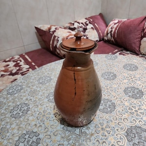 Puede incluir: Una jarra de terracota marrón con cuerpo redondeado, cuello estrecho y una tapa circular a juego con un pequeño pomo. La superficie presenta un acabado liso con sutiles variaciones de color que van desde el bronceado claro al marrón oscuro.