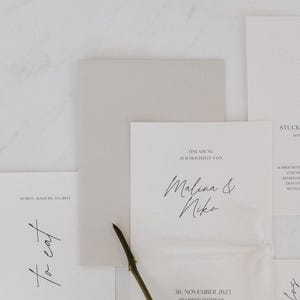 Könnte beinhalten: Eine Ansammlung eleganter Hochzeitspapeterie, darunter Einladungskarten und Menükarten, auf einer Marmoroberfläche arrangiert. Die Einladungskarte zeigt die Namen "Malina & Niko" in Schreibschrift sowie das Datum "30. November 2023". Ein Zweig fügt ein natürliches Element hinzu.