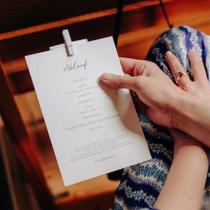 Könnte beinhalten: Eine weiße Karte mit dem Wort "Ablauf" oben, gehalten von einer Holzklammer. Die Karte listet mehrere deutsche Wörter auf, darunter "Einzug" und "Trauung". Die Person, die die Karte hält, trägt einen Ring und ein blau-weiß gemustertes Kleidungsstück.