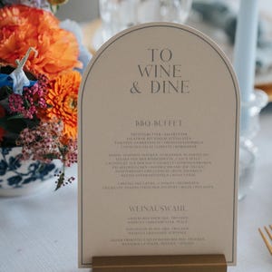 Könnte beinhalten: Eine bogenförmige, beige Menükarte mit dunkelgrauem Text, der TO WINE & DINE, BBQ-BUFFET und WEINAUSWAHL lautet, gehalten von einem rechteckigen, goldfarbenen Metallständer. Die Karte hat einen dünnen, dunkelgrauen Rand entlang der Oberkante.