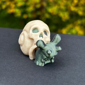 May include: A beige skull figurine with a green metallic mouse figurine. The skull has eye and nose sockets. The mouse is positioned in front of the skull. The background is blurred with green and yellow tones.