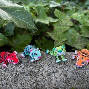 Puede incluir: Cuatro pequeñas figuritas de tigre, cada una con un esquema de color diferente. Los tigres están en una pose juguetona, con la cabeza inclinada y la cola enroscada. Las figuritas están hechas de un material plástico duradero y son perfectas para agregar un toque de fantasía a cualquier habitación.