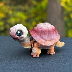 Op de afbeelding: Een klein, 3D-geprint speelgoed schildpad met een roze, oranje en gele schild. De schildpad heeft grote, zwarte ogen en een wit gezicht.