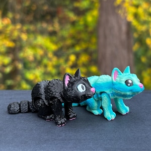 Op de afbeelding: Twee kleine, 3D-geprinte kattenfiguren. Eén is zwart met roze oren en een wit oog. De andere is blauw en groen met roze oren en witte ogen.