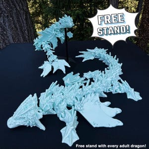 May include: A light blue, articulated dragon model, disassembled into multiple pieces. The dragon is posed in a standing position with its wings spread. The image includes the text "FREE STAND!" in a speech bubble. The caption below the image reads "Free stand with every adult dragon!"