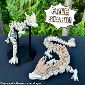 Puede incluir: Dos figuras de dragón blancas y detalladas. Un dragón está de pie sobre un soporte negro. El otro dragón está acostado. La imagen incluye el texto "FREE STAND!" en un bocadillo.