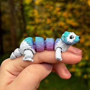 Puede incluir: Un juguete articulado y colorido con forma de animal, parecido a una comadreja o un hurón, sostenido en la mano de una persona. El juguete presenta un degradado de azul, morado y blanco. La cabeza y la cola son blancas con detalles azules. El juguete es segmentado.