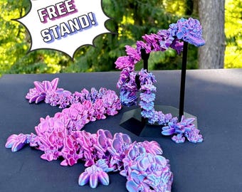 May include: Two iridescent purple and pink dragon figurines with a free stand. The dragon on the left is laying down and the dragon on the right is curled up. The text "FREE STAND!" is in a speech bubble in the top left corner of the image.
