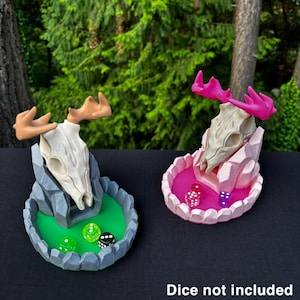 May include: Two 3D printed dice towers shaped like a deer skull with antlers. The towers are made of a light gray plastic and have a green and pink base. The towers are sitting on a black surface. Dice not included.