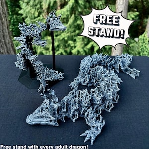 May include: Two 3D printed dragon sculptures, one standing on a black stand and the other lying down. Both dragons are made of a white filament with a textured surface. The image includes the text "FREE STAND!" in a speech bubble.