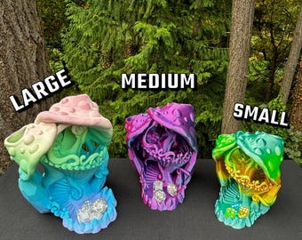 Puede incluir: Tres esculturas de calaveras de fantasía coloridas en diferentes tamaños: grande, mediano y pequeño. La escultura grande es de color pastel, la mediana es morada y rosa, y la pequeña es verde y amarilla. El texto "Dice not included" está en la parte inferior.