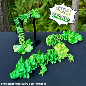 Op de afbeelding: Vier groene klaverdrakenfiguren, waarvan er één op een zwarte standaard staat. De draken zijn gemaakt van 3D-geprint plastic en hebben een textuur. De afbeelding bevat de tekst "FREE STAND!" in een gele spraakballon.