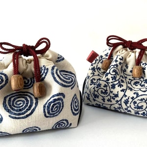 May include: Two small drawstring bags. One features a cream fabric with blue spirals, the other a floral design. Both have red drawstrings and wooden beads. The bags are made of fabric.