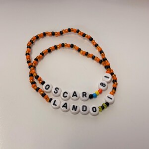 May include: Two beaded bracelets made of small orange and black glass beads. One bracelet features white round beads spelling OSCAR 81 with a blue and yellow bead accent. The second bracelet features white round beads spelling LANDO 1 with a green bead.