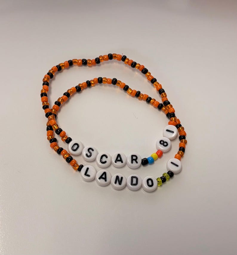 May include: Two beaded bracelets made of small orange and black glass beads. One bracelet features white round beads spelling OSCAR 81 with a blue and yellow bead accent. The second bracelet features white round beads spelling LANDO 1 with a green bead.