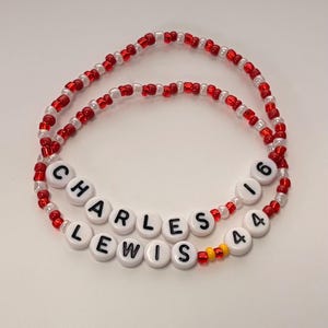 May include: Two beaded bracelets made of small red and white glass beads. One bracelet features white round beads with black letters spelling CHARLES 16, while the other features white round beads with black letters spelling LEWIS 44 with two yellow beads.