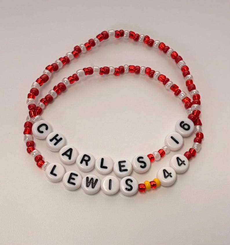 May include: Two beaded bracelets made of small red and white glass beads. One bracelet features white round beads with black letters spelling CHARLES 16, while the other features white round beads with black letters spelling LEWIS 44 with two yellow beads.