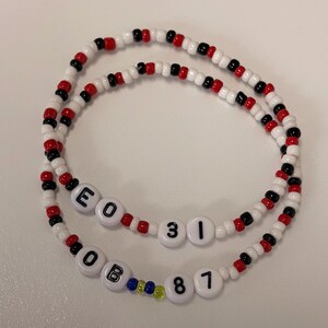 May include: Two beaded bracelets featuring a repeating pattern of white, red, and black seed beads. One bracelet includes white letter and number beads spelling E O 3 1, while the other features white beads spelling O B 8 7 with small blue and yellow accent beads.